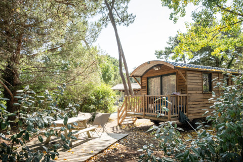 Cabaña Roulotte de madera entre árboles, con terraza, sillas de jardín y banco de picnic al sol.