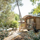 Cabaña Roulotte de madera entre árboles, con terraza, sillas de jardín y banco de picnic al sol.