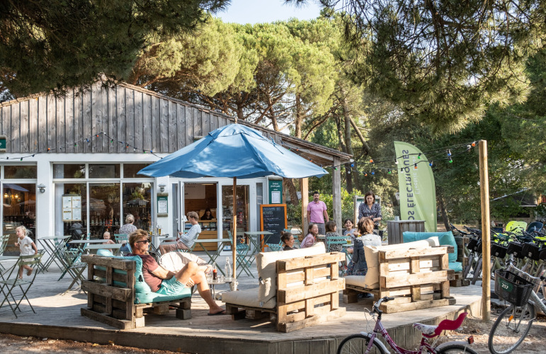 Buitenterras met gezinnen, houten zitbanken en fietsen bij Huttopia Chardons Bleus, Ile de Ré, onder de bomen.