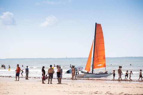Familias disfrutan de un día soleado en la playa cerca de un velero, con niños jugando junto al agua.