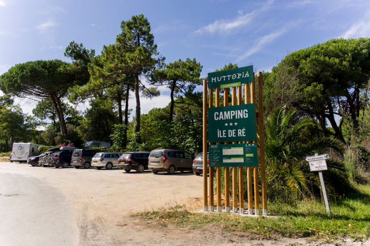 Ingang van Huttopia Chardons Bleus - Île de Ré camping met geparkeerde auto’s en pijnbomen in Nouvelle-Aquitaine.
