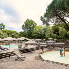 Espace piscine extérieur avec transats, parasols et enfants jouant, entouré d’arbres verdoyants.