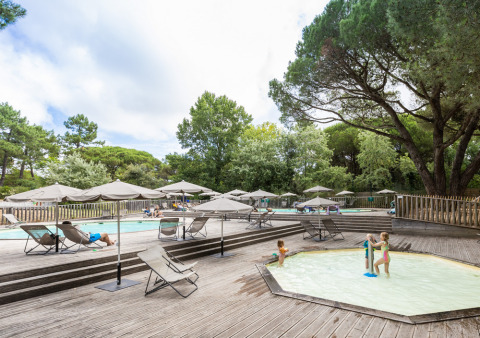 Zona de piscina al aire libre con tumbonas, sombrillas y niños jugando, rodeada de árboles verdes.