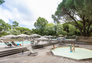 Udendørs swimmingpoolområde med liggestole, parasoller og børn der leger, omgivet af grønne træer.