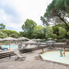 Zona de piscina al aire libre con tumbonas, sombrillas y niños jugando, rodeada de árboles verdes.