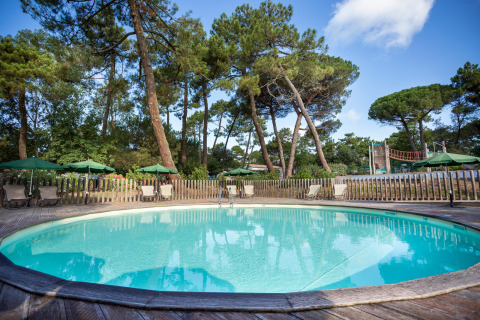 Round swimming pool with deck chairs and green umbrellas, surrounded by pine trees at Huttopia Chardons Bleus, France.