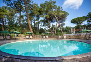 Piscina redonda con tumbonas y sombrillas verdes rodeada de pinos en Huttopia Chardons Bleus, Nueva Aquitania, Francia.