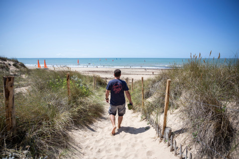 Hombre camina descalzo por un sendero de arena hacia la playa en Huttopia Chardons Bleus - Ile de Ré, Francia.