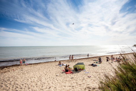 Playa en parque vacacional con glamping; personas descansan cerca de una tienda y disfrutan del mar soleado.