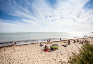 Vakantieparkstrand met glamping; mensen genieten van zon, zee en tent op het zand.