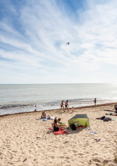 Playa en parque vacacional con glamping; personas descansan cerca de una tienda y disfrutan del mar soleado.
