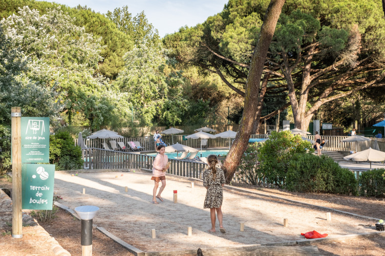 Children play boules on a sandy court at Huttopia Chardons Bleus - Ile de Ré holiday park surrounded by trees.