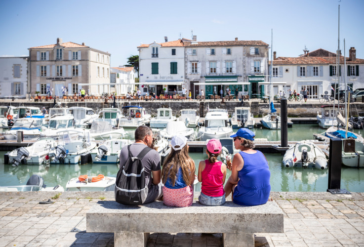 Family sits on a bench by the harbor in Sainte-Marie-de-Ré, France, overlooking boats and seaside buildings.