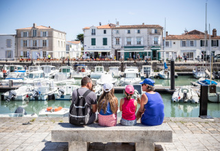 Family sits on a bench by the harbor in Sainte-Marie-de-Ré, France, overlooking boats and seaside buildings.