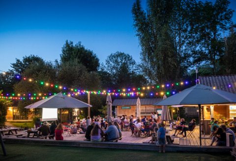 Gezellig terras bij Huttopia Beaulieu sur Dordogne met mensen onder kleurrijke lichtjes op een zomeravond.