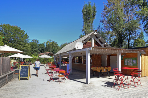Terraza exterior en Huttopia Beaulieu sur Dordogne con mesas rojas, sombrillas y vegetación bajo un cielo despejado.