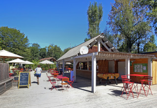 Udendørs caféområde i Huttopia Beaulieu sur Dordogne med røde borde, parasoller og grønne træer under blå himmel.