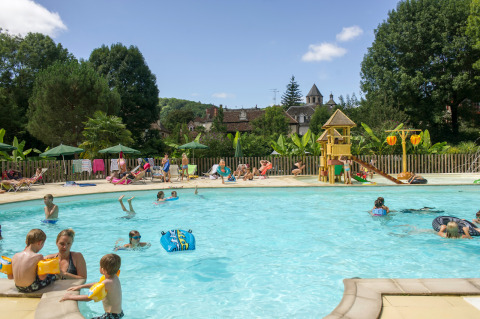 Familias se divierten nadando y descansando junto a la piscina y la zona de juegos de Huttopia Beaulieu sur Dordogne.