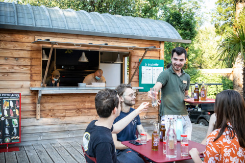 Scène de café en plein air à Huttopia Beaulieu sur Dordogne avec serveurs, clients et cabanon bois.