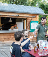 Außencafé bei Huttopia Beaulieu sur Dordogne mit Gästen, Kellner und Holzhütte im Grünen.