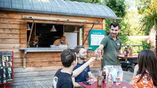 Außencafé bei Huttopia Beaulieu sur Dordogne mit Gästen, Kellner und Holzhütte im Grünen.
