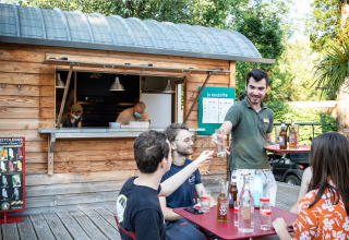 Udendørs café ved Huttopia Beaulieu sur Dordogne med gæster, tjener og træhytte. Sommerstemning.