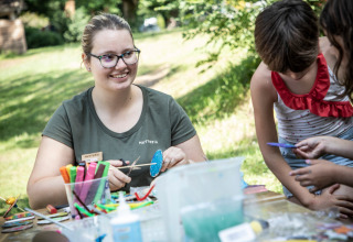 Donna sorridente fa lavoretti con bambini all’aperto a Huttopia Beaulieu sur Dordogne, nel parco vacanze in Occitania.