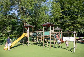 Legeplads med børn, der leger på rutsjebane og gynger under grønne træer i ferieparken Huttopia Beaulieu.