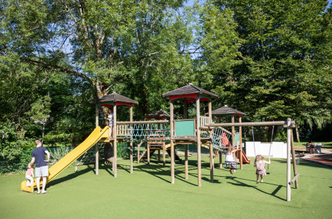Bambini che giocano su scivolo e altalene nel parco giochi di Huttopia Beaulieu sur Dordogne, tra alberi verdi.