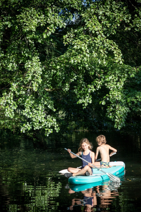 Due bambini remano su una tavola gonfiabile sotto alberi verdi a Huttopia Beaulieu sur Dordogne, Francia.