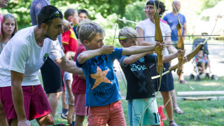 Kinder üben Bogenschießen im Freien unter Anleitung eines Erwachsenen im Huttopia Beaulieu sur Dordogne.