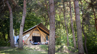 Twee personen relaxen voor een Trappeur safaritent, omringd door hoge bomen en groene natuur in het bos.