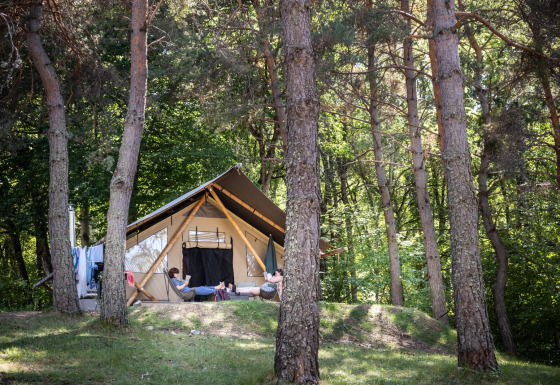 Twee personen relaxen voor een Trappeur safaritent, omringd door hoge bomen en groene natuur in het bos.