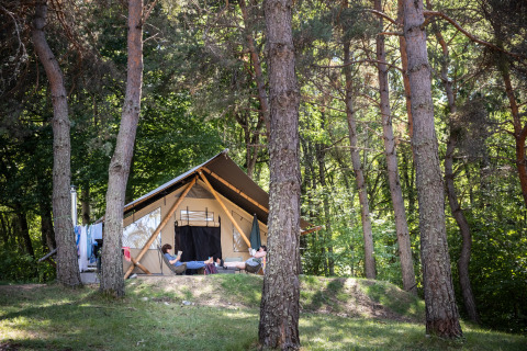 Two people relax in front of a Trappeur safari tent, surrounded by tall trees and lush green forest.