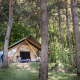 Dos personas se relajan frente a una tienda safari Trappeur, rodeadas de altos árboles y naturaleza verde.