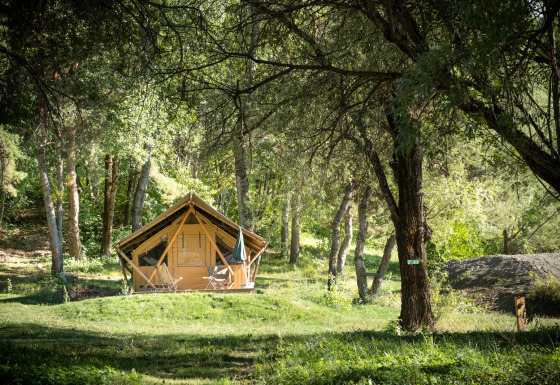 Una tenda safari chiamata Canadienne Tent II si trova in una radura verde, circondata da alberi e con sedie fuori.