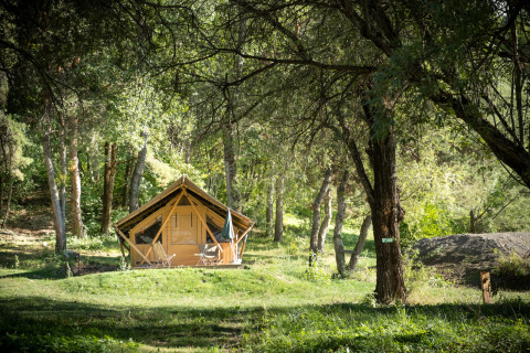 A safari tent called Canadienne Tent II is set in a green forest clearing with chairs outside and dappled sunlight.