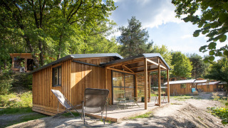 Cabaña de madera Chalet Evasion con terraza techada, sillas y entorno natural lleno de árboles y luz solar.