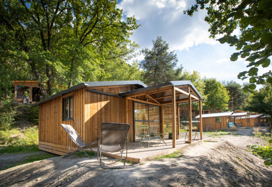 Cabaña de madera Chalet Evasion con terraza techada, sillas y entorno natural lleno de árboles y luz solar.