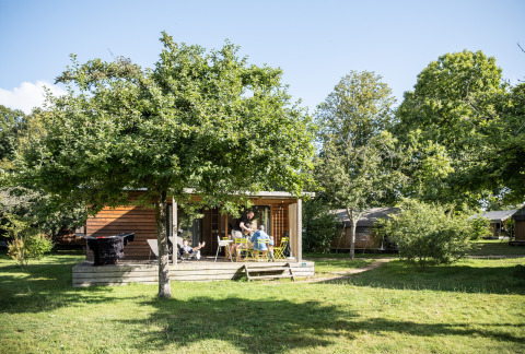 Una acogedora cabaña de madera con terraza y muebles de exterior bajo árboles en Chalet Portland, Huttopia Lac de Serre Ponçon.