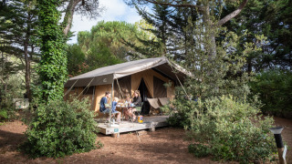 Familie sidder ved et bord foran en safari-telt Toile&Bois tent Sweet i grønne omgivelser.