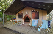 Tienda safari sobre plataforma de madera con sillas, mesa y sillones en un campamento arbolado.