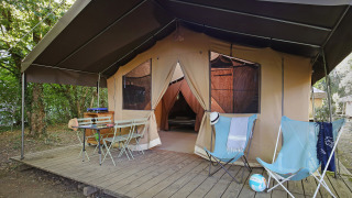 Tienda safari sobre plataforma de madera con sillas, mesa y sillones en un campamento arbolado.