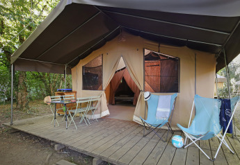 Tienda safari sobre plataforma de madera con sillas, mesa y sillones en un campamento arbolado.