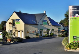 Entrance to Camping Saint Michel holiday park in Normandy, France, showing buildings and lush landscaping.