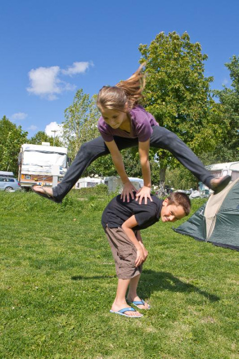 Two kids play leapfrog on green grass under a blue sky at Camping Seasonova Saint Michel in Normandy, France.