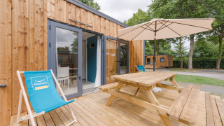 Outdoor deck at Prestige Cottage with wooden picnic table, parasol, and loungers near glass doors.