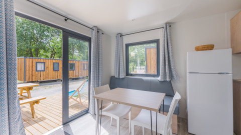 Interior de una cabaña moderna con mesa de comedor, sillas, nevera y vista a terraza de madera y otras cabañas.