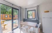 Interior de una cabaña moderna con mesa de comedor, sillas, nevera y vista a terraza de madera y otras cabañas.