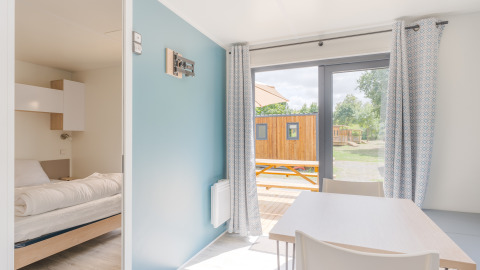 Bright lodge interior with cozy bedroom, dining table, large windows, curtains, and view of deck outside.